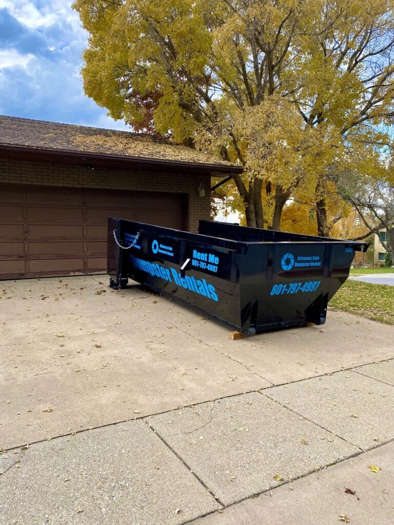 A black 15 yard dumpster rental container parked on a residential driveway in front of a house.