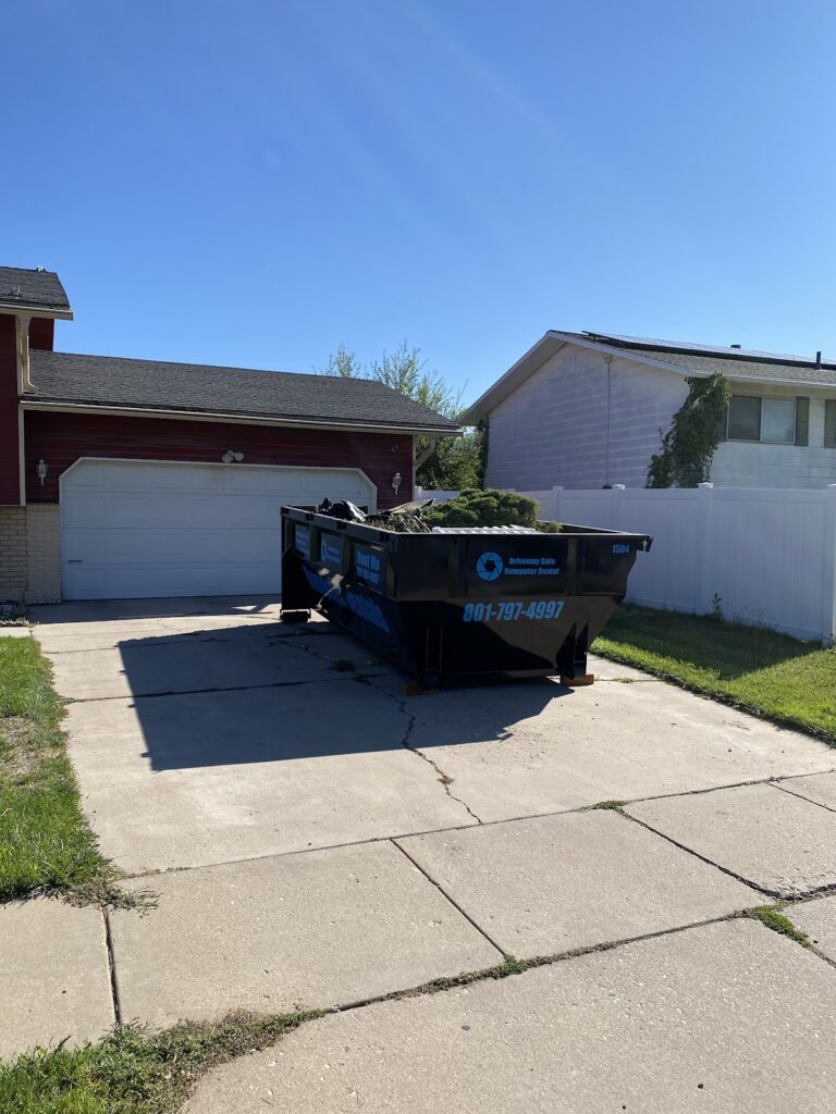 A black roll off dumpster rental parked on a residential driveway in front of a house, featuring the text “Driveway Safe Dumpster Rental” and the phone number 801-797-4997.