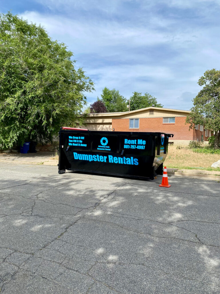 A large black dumpster labeled "Dumpster Rentals" sits on a residential street beside a traffic cone, with trees and a house in the background, highlighting convenient dumpster rental Utah options and affordable dumpster rental pricing.
