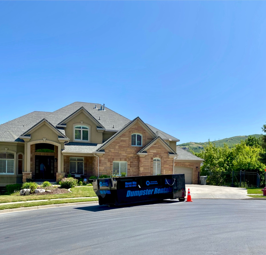 A large house with a stone facade and gray roof in Roy has a dumpster from local Dumpster Rental Services and an orange traffic cone placed on the street in front of it on a sunny day.