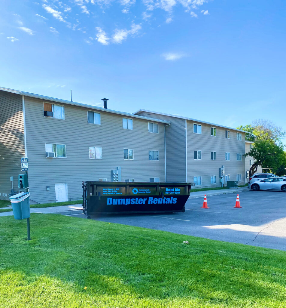 A large dumpster labeled "Dumpster Rentals" sits in a parking lot in front of a beige apartment building, with two orange traffic cones nearby, following roll-off dumpster guidelines for safe placement.