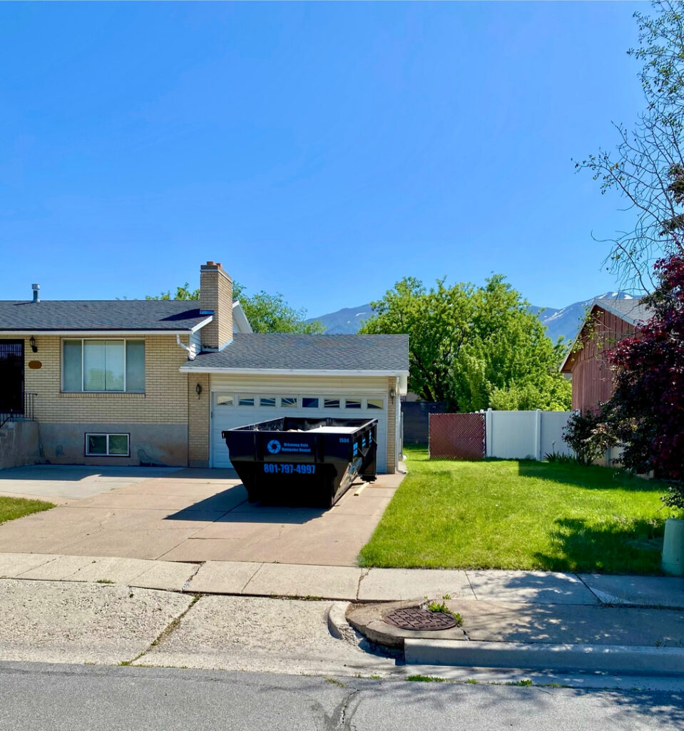 A residential house with a large black Roy Dumpster placed in the driveway, surrounded by a lawn, trees, and clear blue sky.