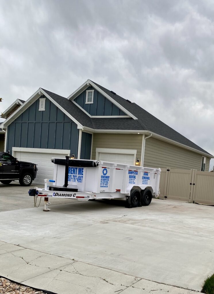 A white dumpster trailer labeled "RENT ME" and a phone number is parked in the driveway of a two-story house with a garage on a cloudy day in Utah. This driveway safe option makes clean-up easy while offering affordable dumpster rental prices.
