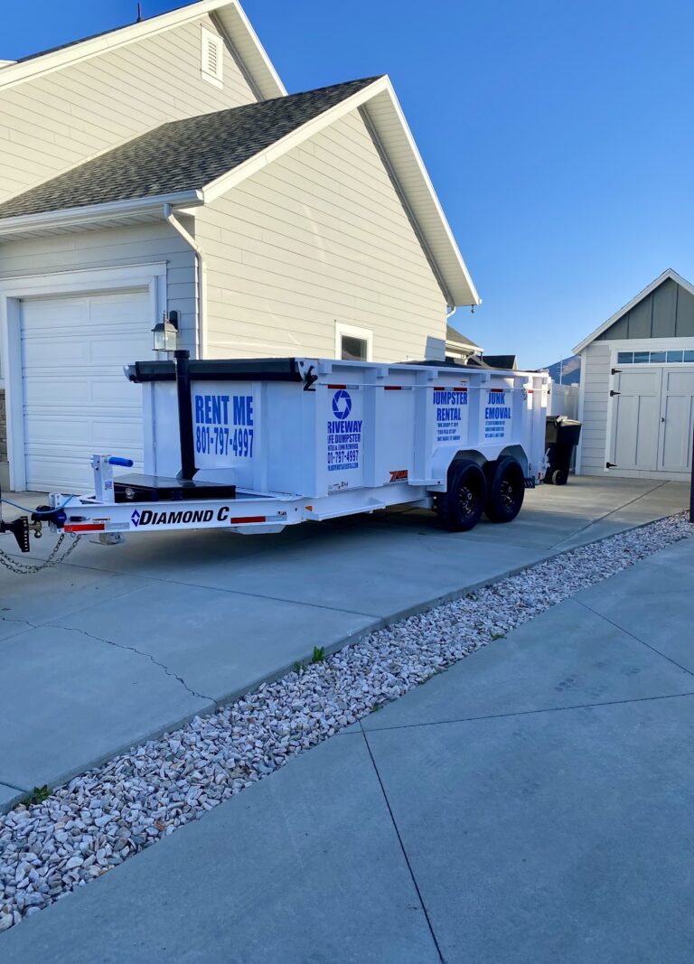 White dumpster trailer with company logos and a "RENT ME" sign parked on a driveway beside a light-colored house and a garage, with landscaping rocks along the driveway edge. This Utah-based dumpster rental offers convenient, driveway safe solutions at competitive dumpster rental prices.