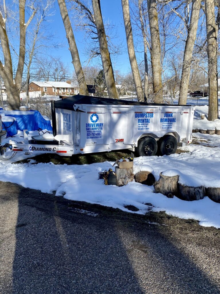 A white dumpster rental trailer with blue signage is parked on a snowy ground near trees and cut tree stumps on a sunny day, offering Utah residents a driveway safe solution with competitive dumpster rental prices.