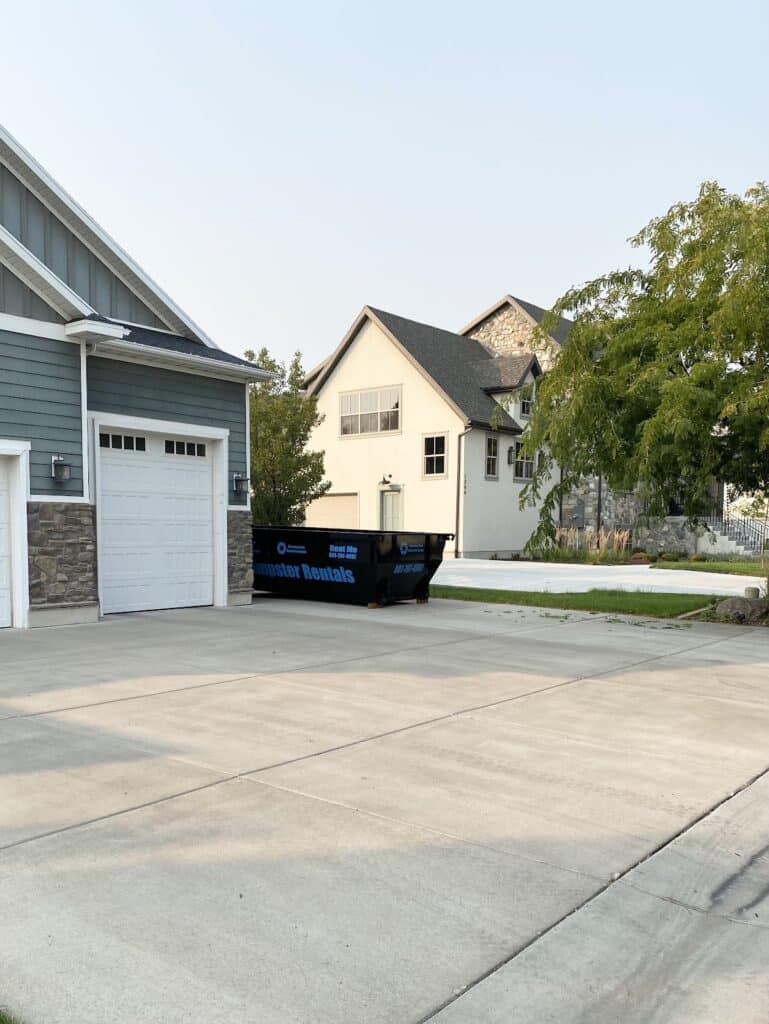 A black roll-off dumpster labeled “Neighbor Rentals” sits on the driveway in front of a residential garage, ready to help with neighborhood projects, with houses and trees in the background.