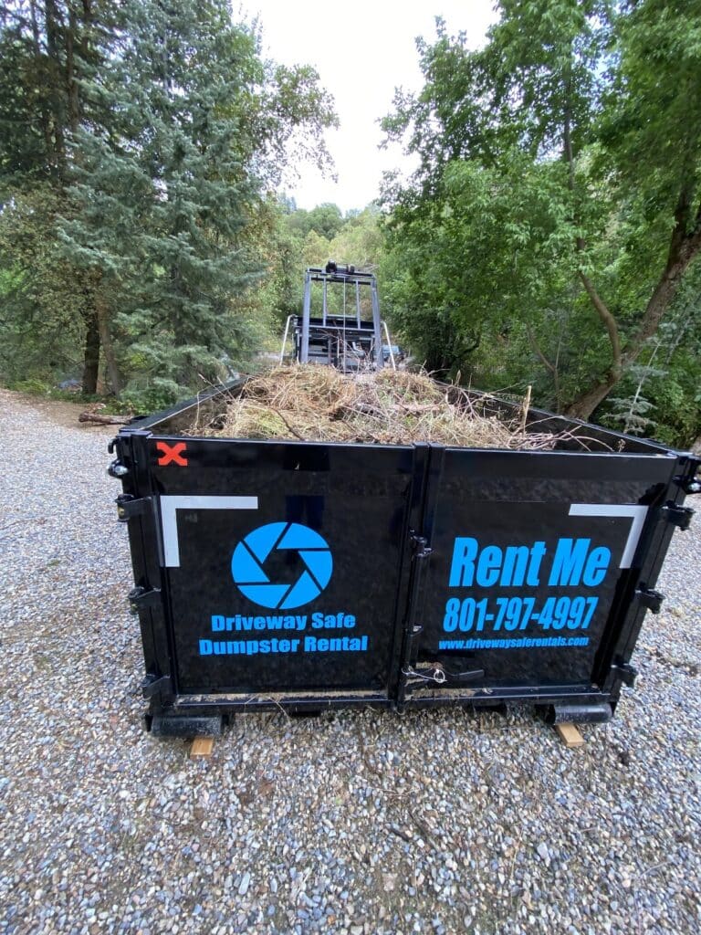 A black dumpster filled with yard debris sits on a gravel driveway, surrounded by trees. The dumpster displays “Driveway Safe Dumpster Rental” and a phone number, making it an ideal choice for neighborhood projects in need of convenient roll-off dumpster rentals.