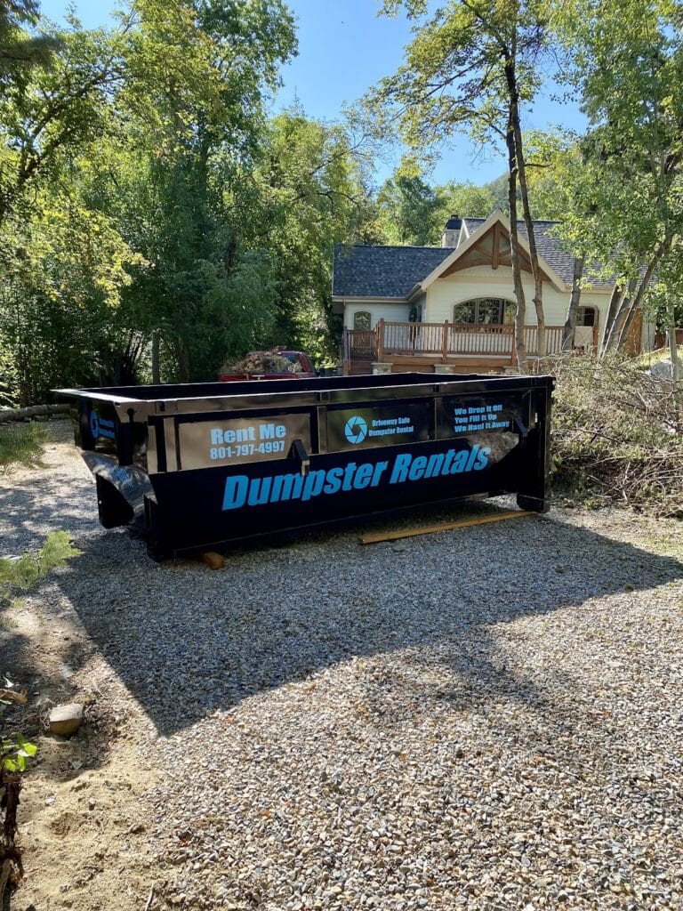 A 20 yard dumpster for rent parked on a gravel driveway in front of a house, featuring the company phone number and rental instructions.