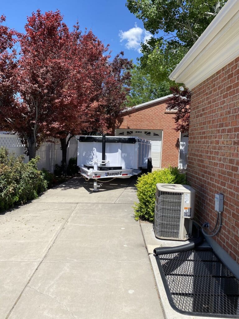 A white trailer—similar in function to driveway safe dumpsters—is parked in a driveway next to a brick house with red-leaved trees and an outdoor air conditioning unit visible.