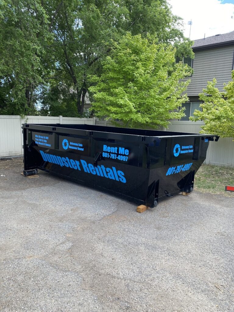 A large black dumpster labeled “Dumpster Rentals” sits on gravel near trees and a house, with rental contact information displayed on the sides. This driveway safe dumpster is an ideal option for those seeking convenient dumpsters for rent to handle their cleanup or renovation projects.