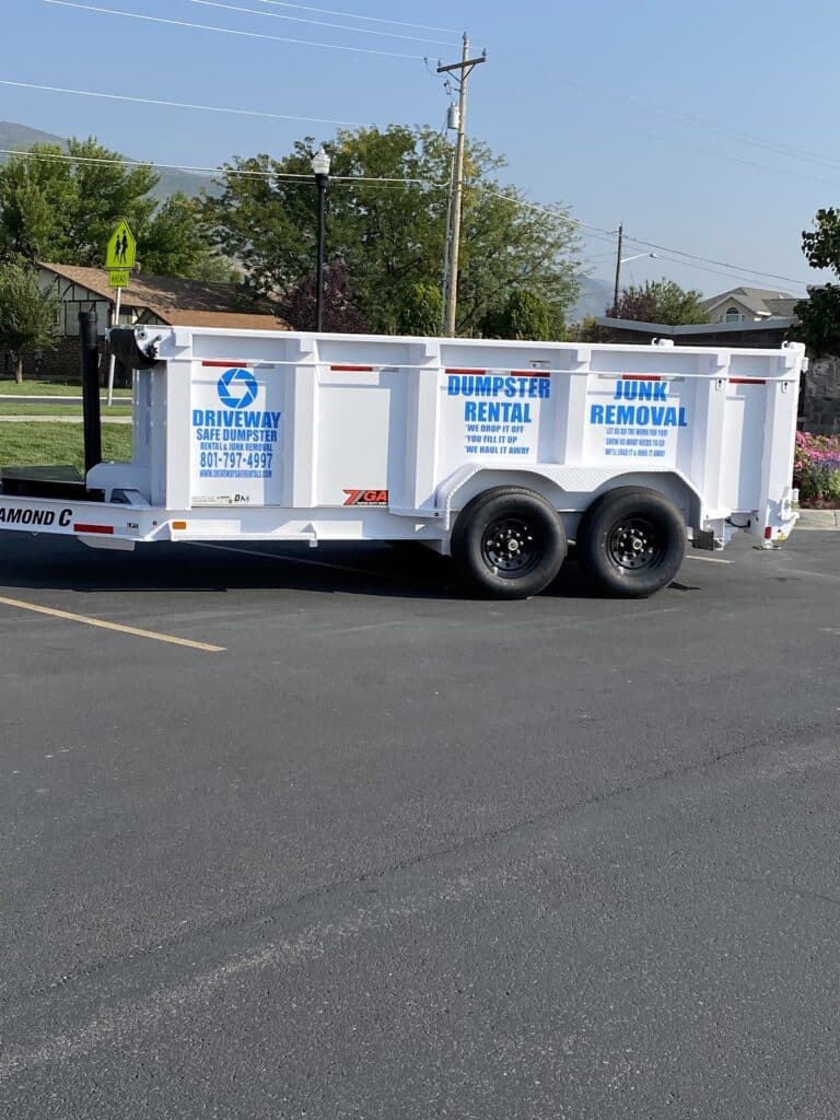 White trailer labeled “Dumpster Rental” and “Junk Removal” is parked on asphalt, featuring a phone number and business logo on the side. Ideal for Neighborhood Projects, this trailer offers convenient Dumpster Rentals solutions. Trees and utility poles are visible in the background.