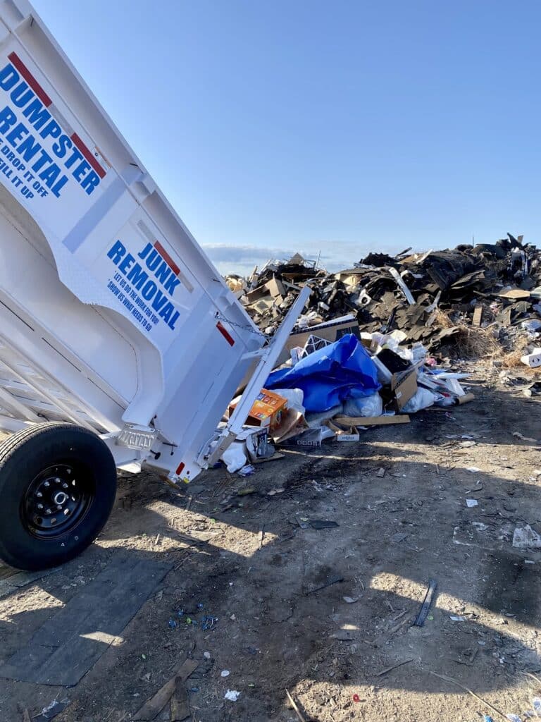 A dumpster trailer labeled “Junk Removal” is unloading debris at a landfill site with a large pile of trash in the background under a clear sky, showcasing one of our convenient Dumpsters for Rent as an efficient solution for large-scale cleanups.