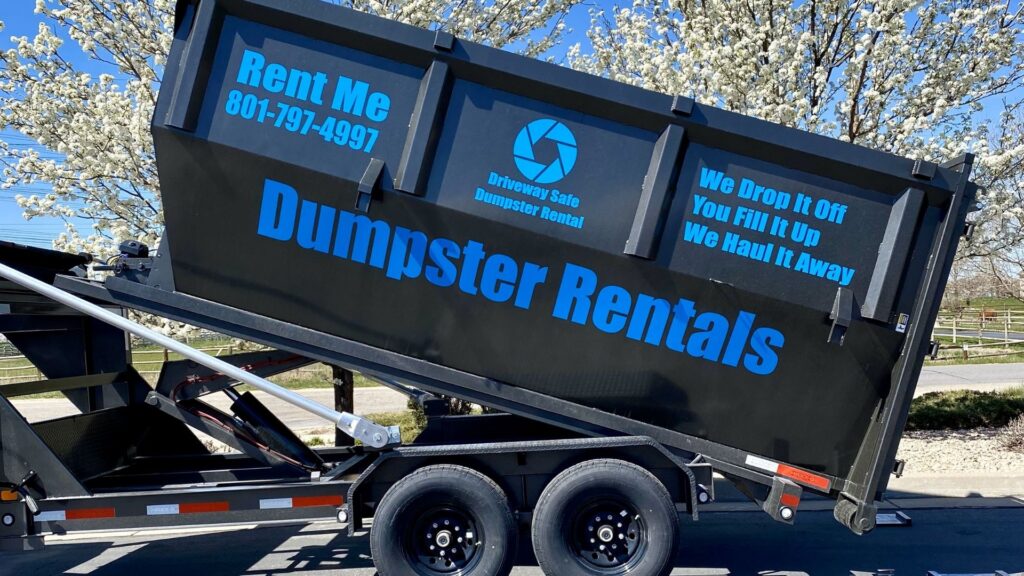 A black roll-off dumpster labeled "Dumpster Rentals" sits on a trailer in Utah, with contact number and service details, parked outdoors near blooming trees—ideal for cleanups, including tires.
