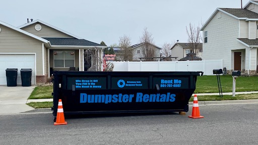 A black dumpster labeled "Dumpster Rentals" sits on a residential street between two orange cones in front of suburban homes.