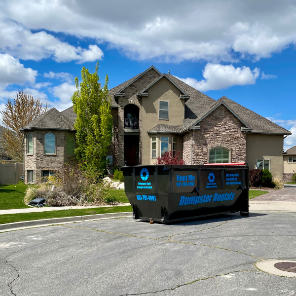 A large residential house with a dumpster rental placed at the edge of the driveway on a suburban street, under a blue sky with scattered clouds—perfect for your next home improvement project.