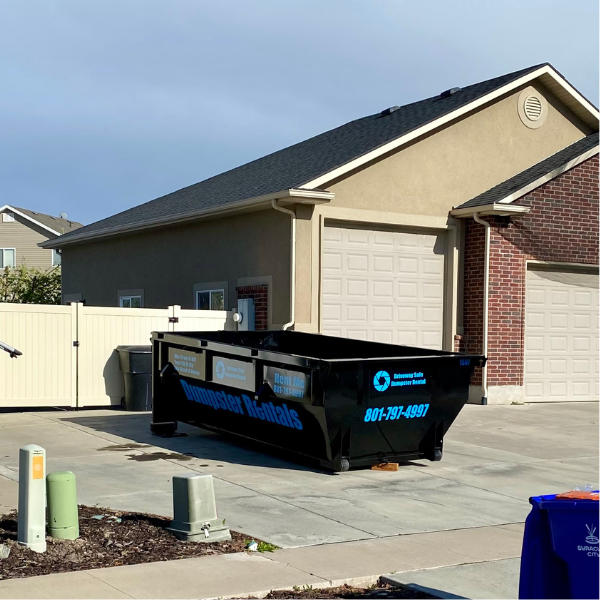 A large black dumpster labeled "Dumpster Rentals" sits on a driveway in front of a beige house with a two-car garage, ready to support your next home improvement project with reliable dumpster rental services.