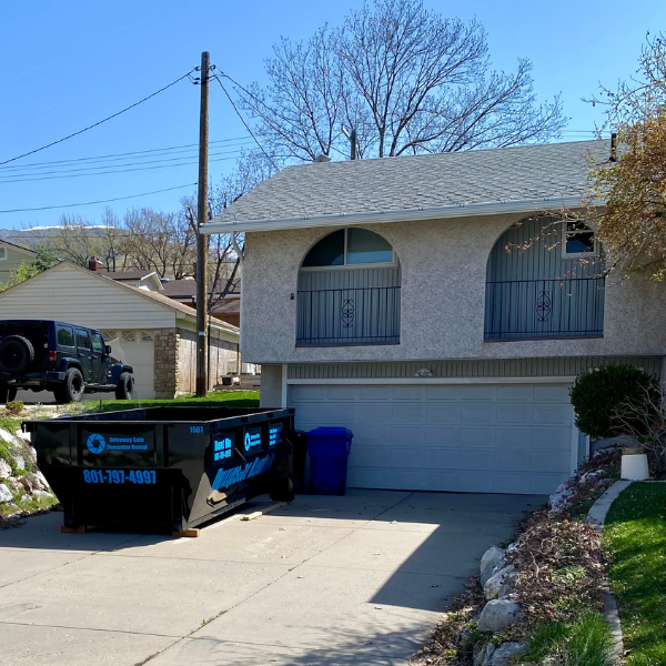 A residential house with a two-car garage has a large black dumpster from a Dumpster Rental service and a blue recycling bin placed on the driveway, making cleanup easy during your home improvement project.