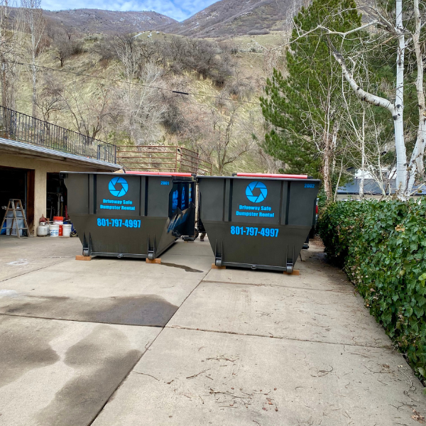 Two large metal dumpsters with blue logos and phone numbers are placed on a concrete driveway near a hillside and some trees, ready for your next home improvement project. Convenient dumpster rental makes cleanup easy.