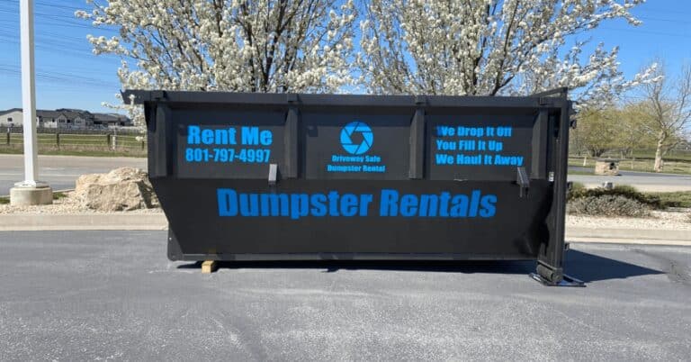 Large black dumpster labeled "Dumpster Rentals" with contact information and rental instructions, placed in a parking lot near blossoming trees—ideal for any home project. Check the dumpster guide for available residential dumpster sizes.