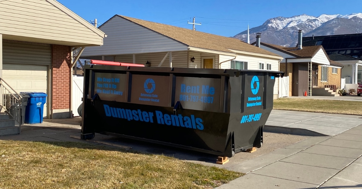 A large black dumpster labeled "Dumpster Rentals" sits in the driveway, perfectly placed to ensure driveway protection, on a sunny day between two houses.