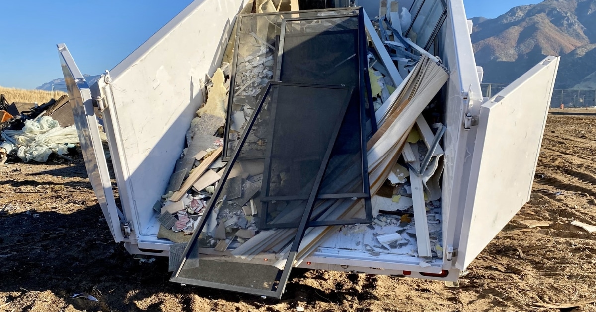 A rented dumpster filled with construction debris, including broken windows and various building materials, sits outdoors against a backdrop of mountains.