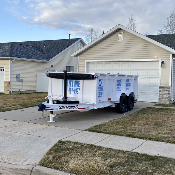 A white 15-yard dumpster rental trailer is parked on a driveway in front of a two-car garage in a suburban neighborhood, displaying company branding and a phone number.
