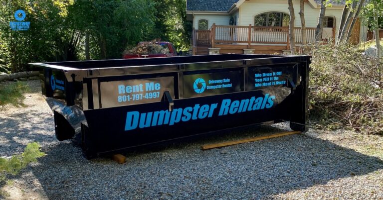 A large black 15-yard dumpster with "Dumpster Rentals" and a phone number printed on the side sits on a gravel driveway in front of a house surrounded by trees.
