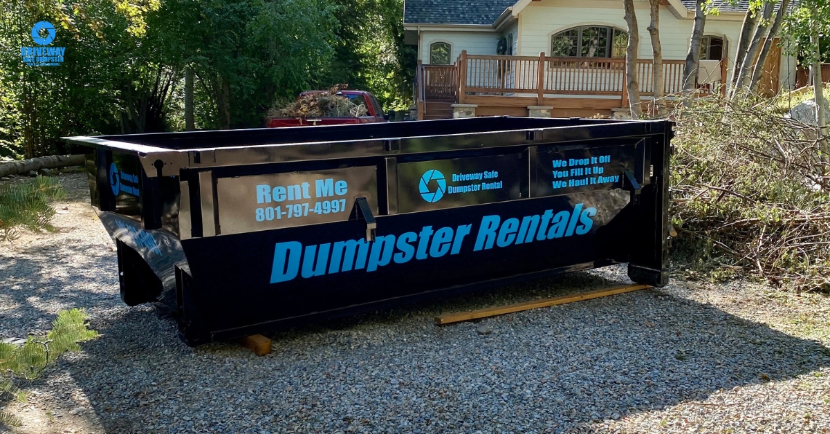 A large black 15-yard dumpster with "Dumpster Rentals" and a phone number printed on the side sits on a gravel driveway in front of a house surrounded by trees.