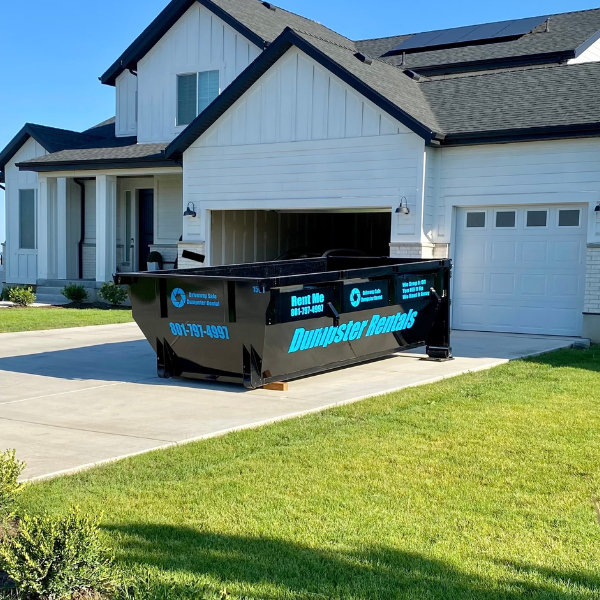 A large black 15-yard dumpster labeled "Dumpster Rentals" sits on the driveway of a modern white house, showcasing the perfect dumpster size for a sunny day project.