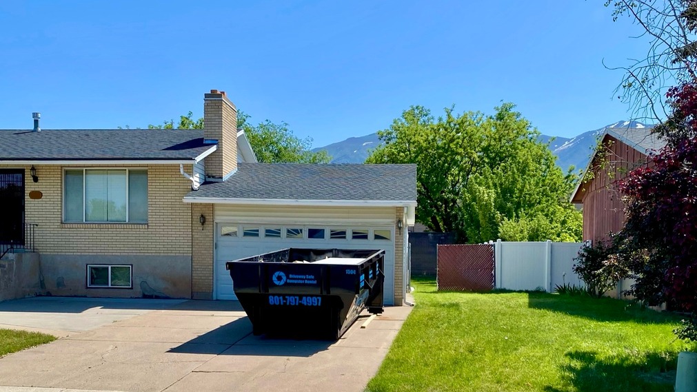 A residential driveway with a black dumpster—provided by a Utah dumpster rental—sits in front of a two-car garage. The house features light brick siding, a green lawn, and scenic trees and mountains in the background.