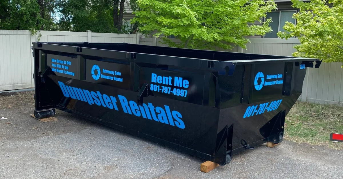 Large black dumpster labeled "Dumpster Rentals" with blue text and contact info, placed on pavement with wooden blocks under one end, near a white fence and green tree—perfect for projects following roll-off dumpster guidelines.