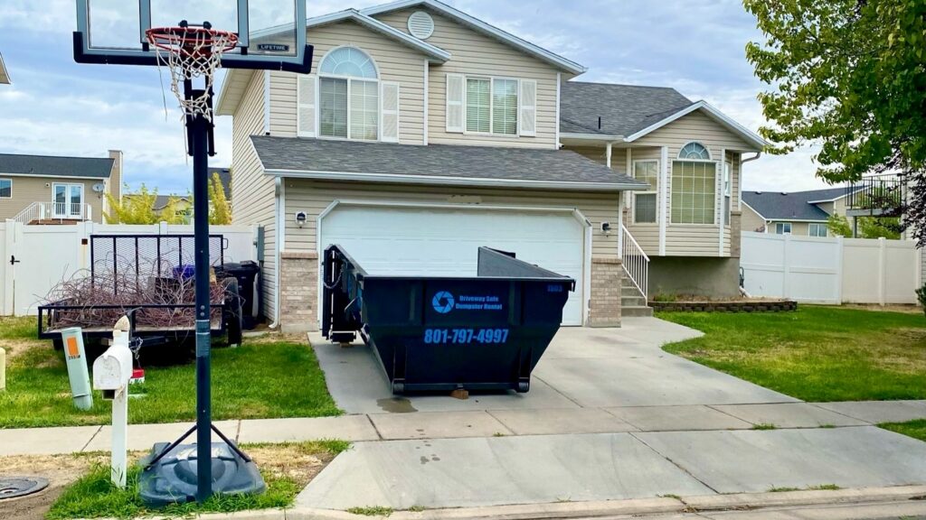 A light beige suburban house with a double garage, a dumpster rental Utah in the driveway, a basketball hoop by the curb, and a white mailbox in front.