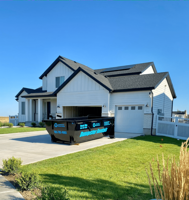 A black residential dumpster rental container parked in the driveway of a white suburban house.