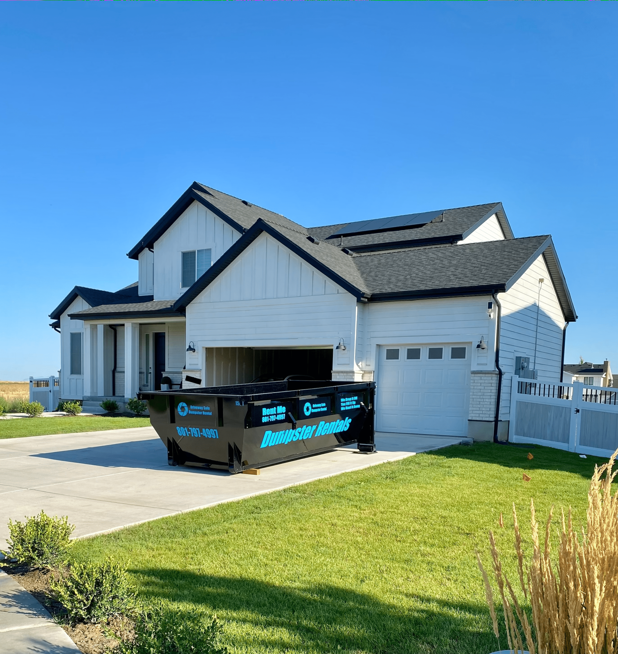 A black residential dumpster rental container parked in the driveway of a white suburban house.