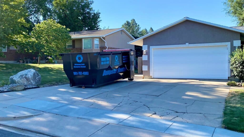 A black dumpster sits on a cracked driveway in front of a two-car garage and a suburban house with green lawns and trees in Davis County, UT, perfect for your next dumpster rental project.