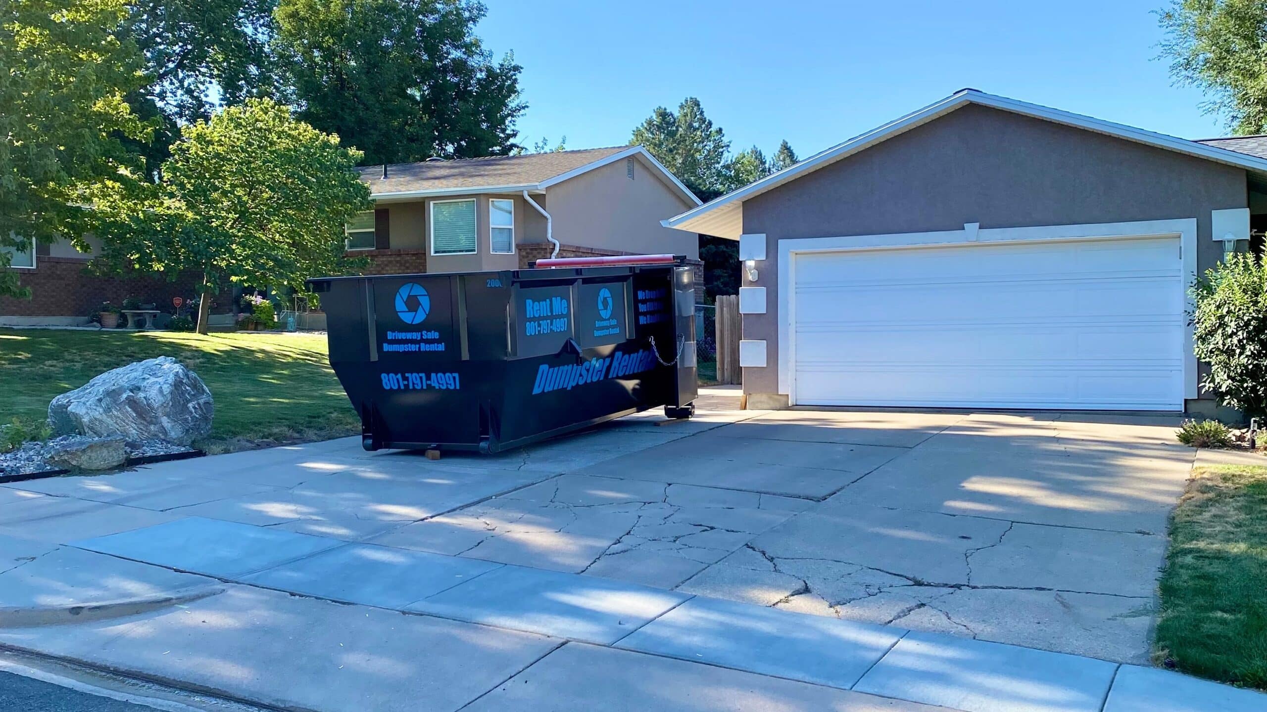 A black dumpster sits on a cracked driveway in front of a two-car garage and a suburban house with green lawns and trees in Davis County, UT, perfect for your next dumpster rental project.