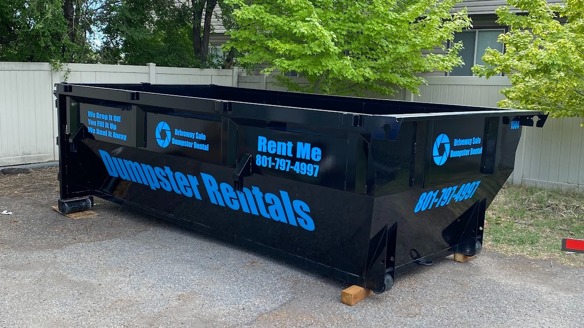 Large black dumpster labeled “Dumpster Rentals” with blue text and a rental phone number, placed on wooden blocks in an outdoor area near trees and a white fence. This driveway-safe dumpster is ideal for property cleanouts, ensuring both convenience and protection for your surfaces as you tackle your cleanup project.