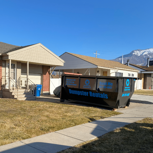 A large black dumpster rental for rent sits in the driveway of a suburban Farmington house, with Northern Utah mountains visible in the background.
