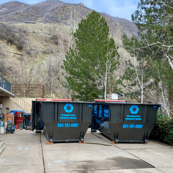 Two large black roll off dumpsters labeled “Driveway Safe Dumpster Rental” sit on a concrete driveway near trees and hills, with cleaning supplies visible on the left—ideal for any dumpster rental period.